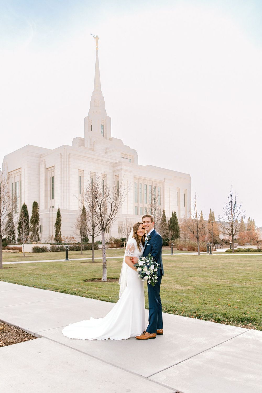 Bride and groom pose on a pathway before a white LDS temple with a golden spire on their wedding day.