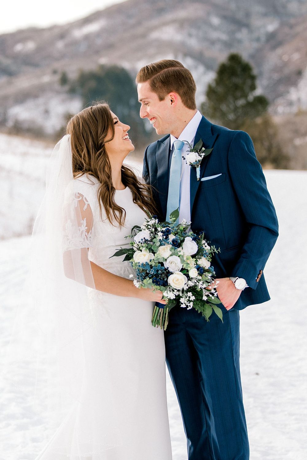 Bride and groom smiling at each other in snowy winter landscape, holding blue and white bouquet, groom in navy suit.