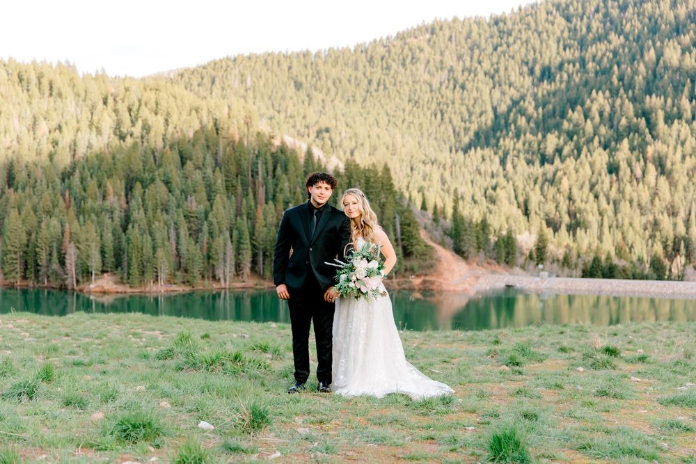 Bride and groom pose by a mountain lake surrounded by pine forests during their outdoor wedding ceremony.