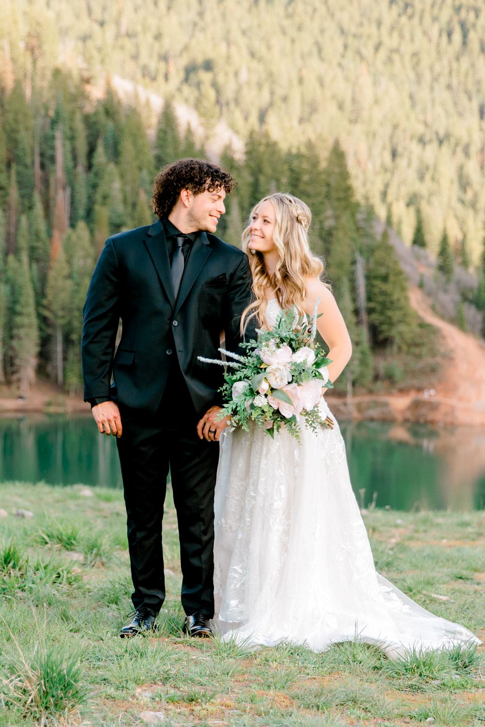 Bride and groom standing by a mountain lake surrounded by pine trees, she holds a blush bouquet.