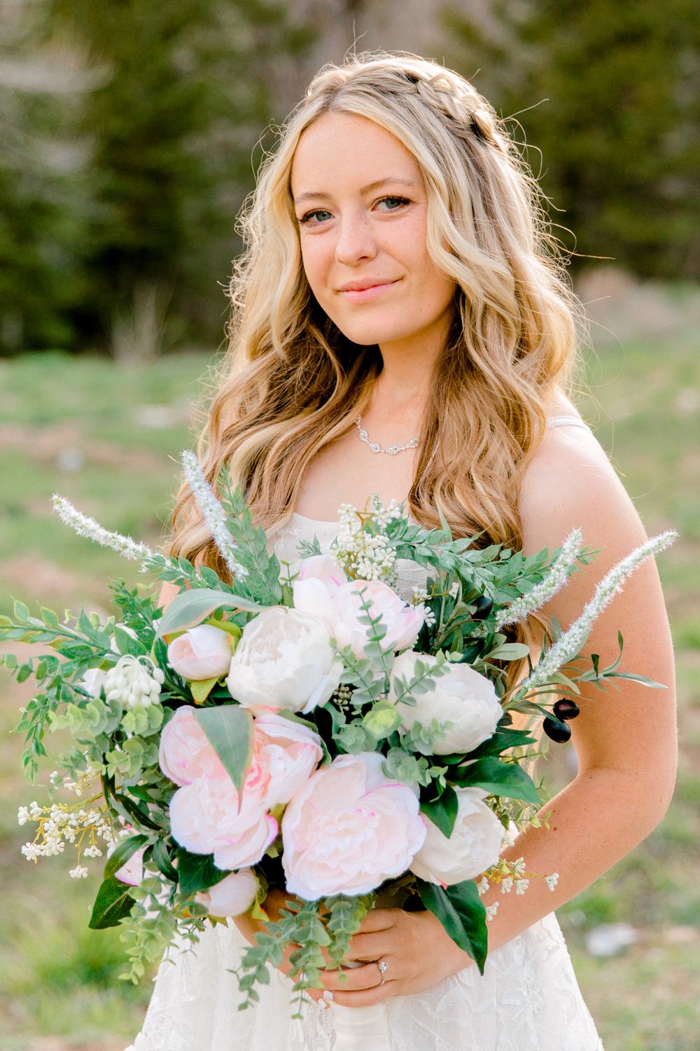 Bride in white dress holding lush bouquet of pink peonies and greenery, smiling outdoors at wedding.