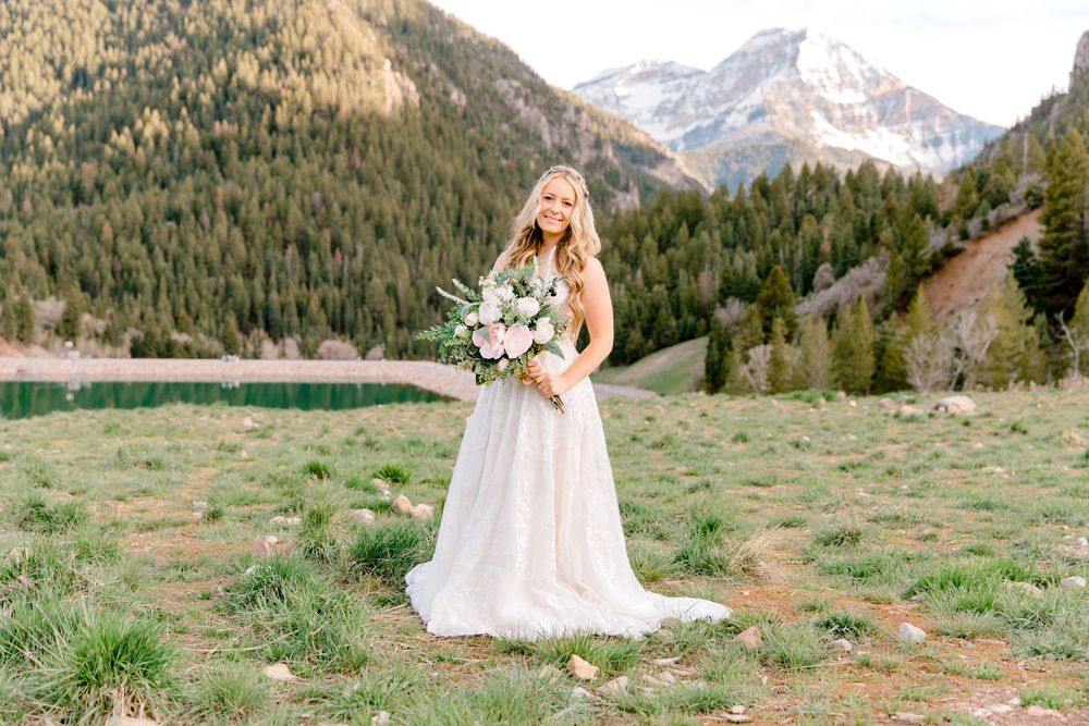 Bride in white gown holding floral bouquet standing in mountain meadow with lake and pine forest backdrop.