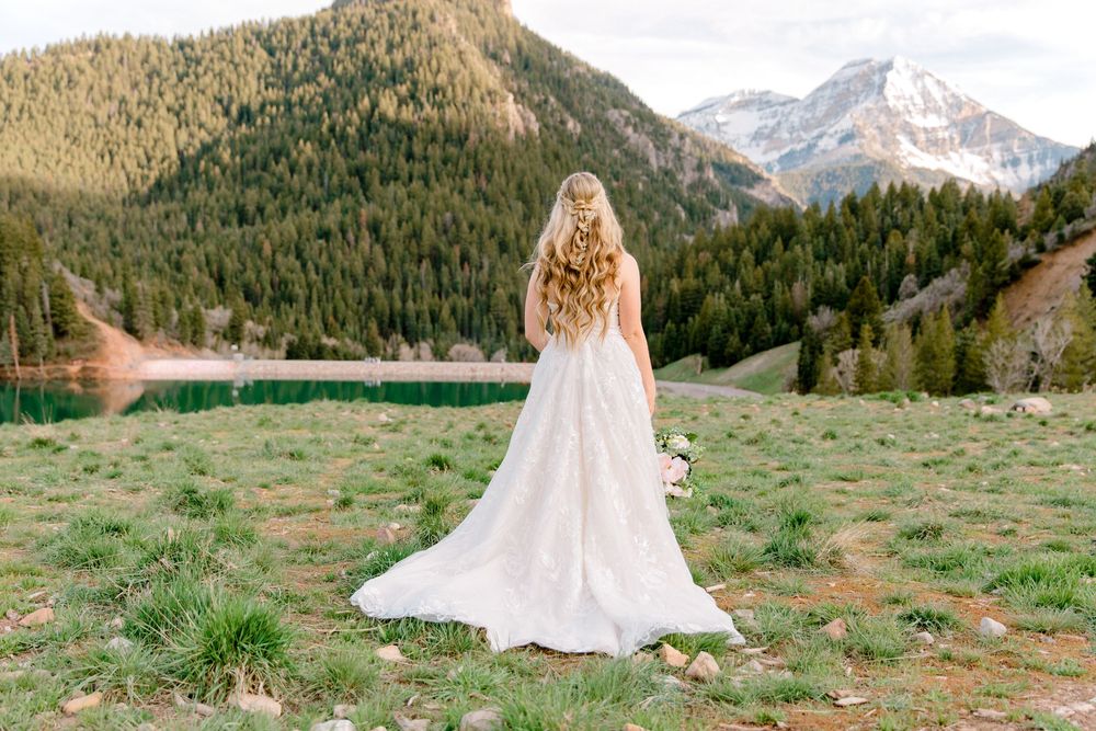 Bride in white gown stands facing mountain lake and pine forest backdrop in scenic outdoor wedding photo.