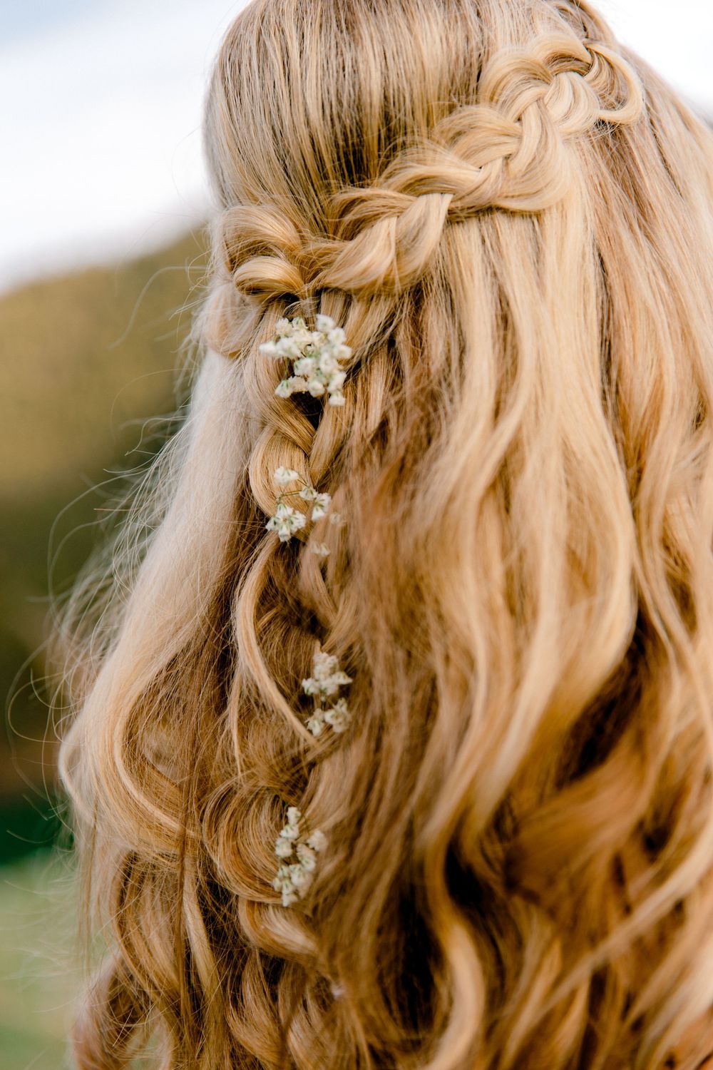 Close-up of bridal half-up hairstyle with braids and small white flowers woven through loose blonde curls.