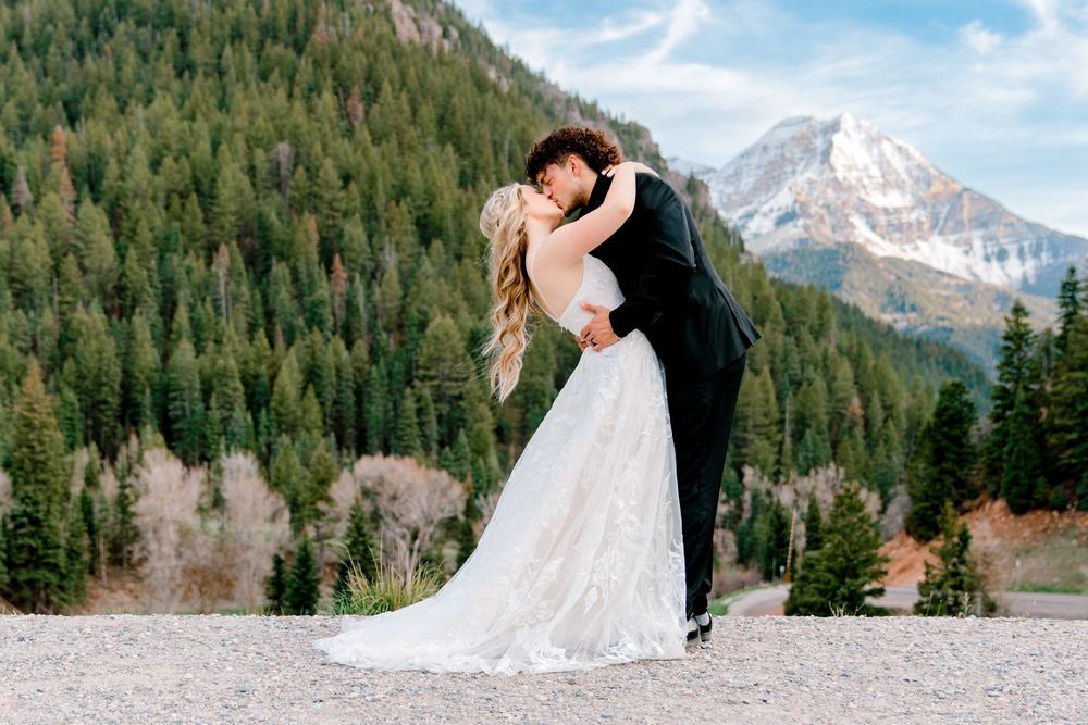 Bride and groom share a romantic dip kiss with snow-capped mountains and pine forest backdrop on their wedding day.