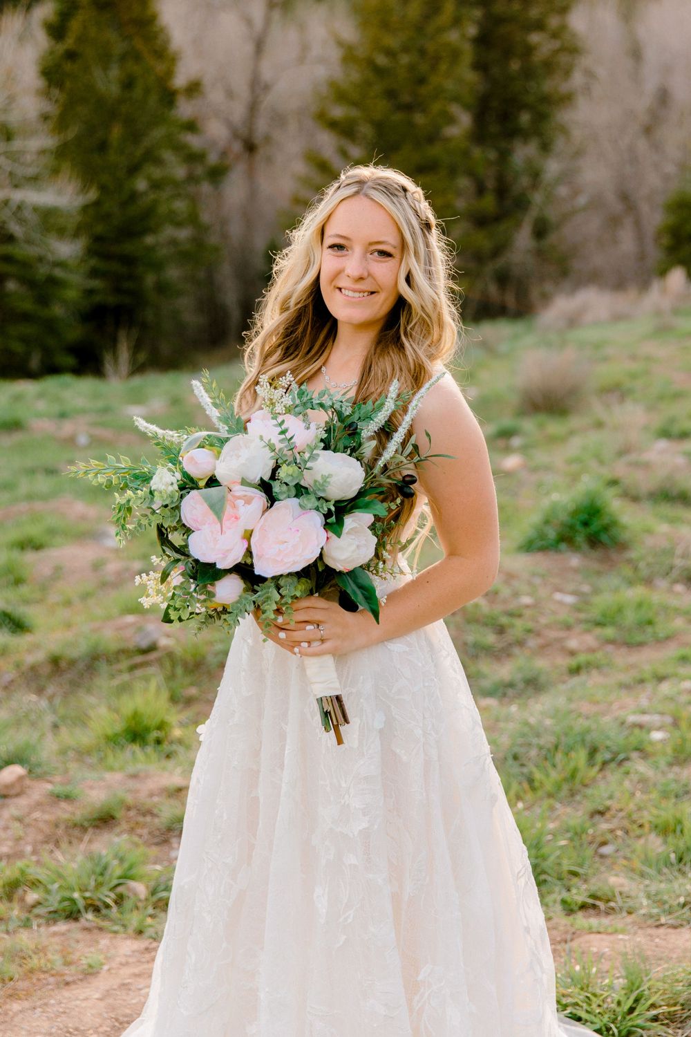 Smiling bride in lace gown holds pink peony bouquet outdoors in a mountain meadow setting.