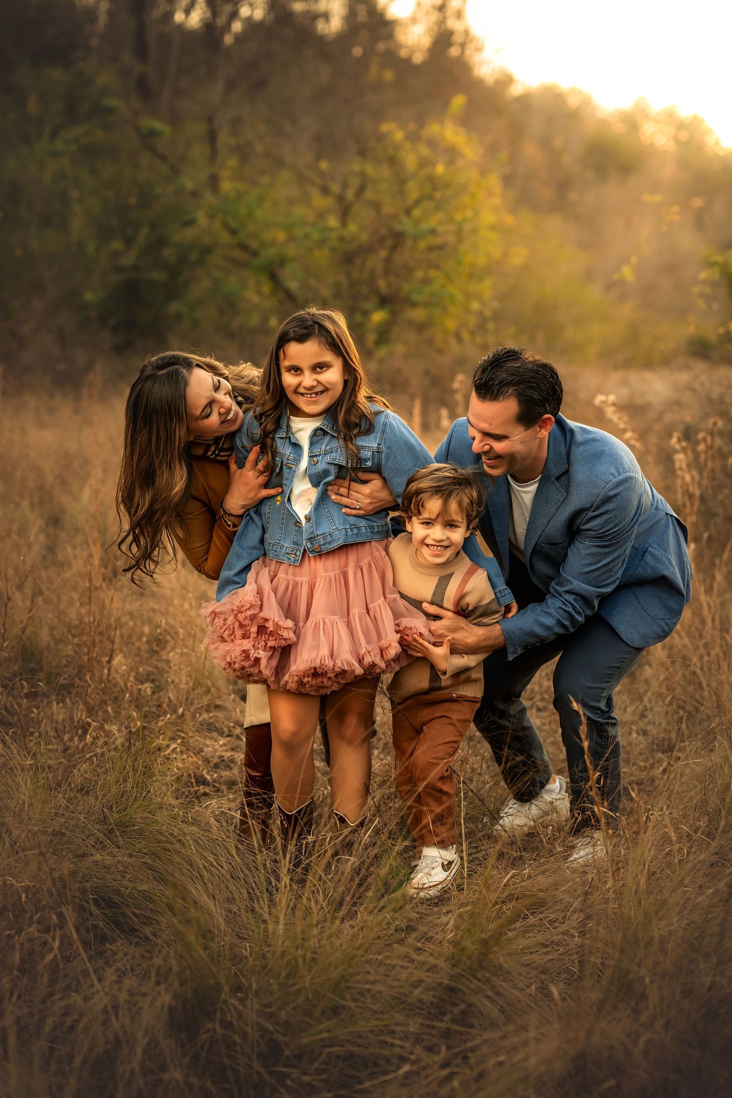 Family of four laughing together in a golden field at sunset, parents playfully tickling two young children outdoors.