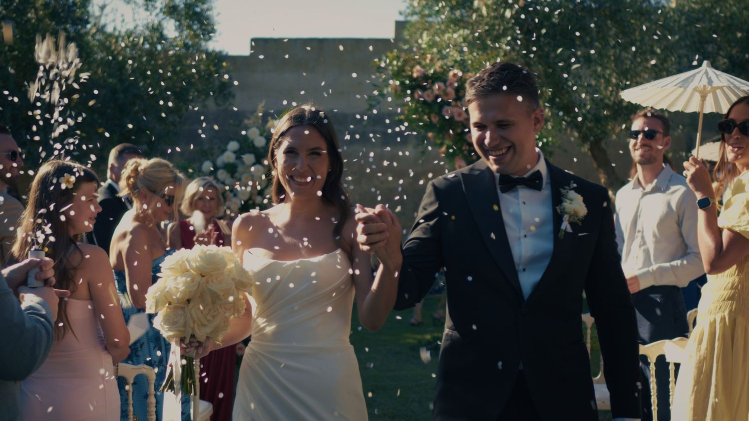 A joyful bride and groom walk through a shower of confetti at their outdoor wedding ceremony surrounded by guests.