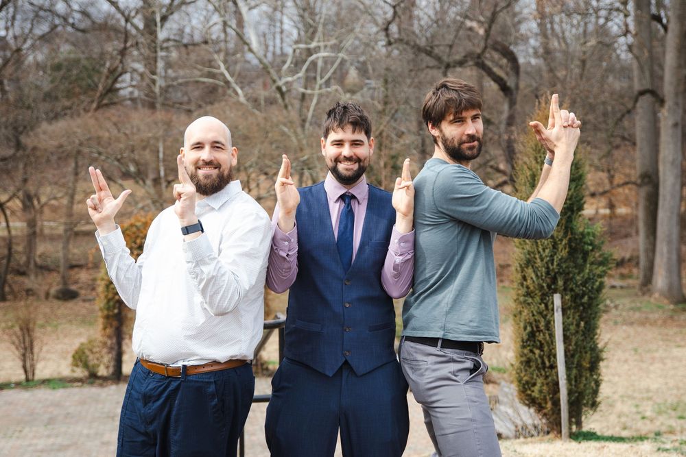 Three men posing outdoors at a wedding, middle man in blue suit flanked by friends pointing fingers up.