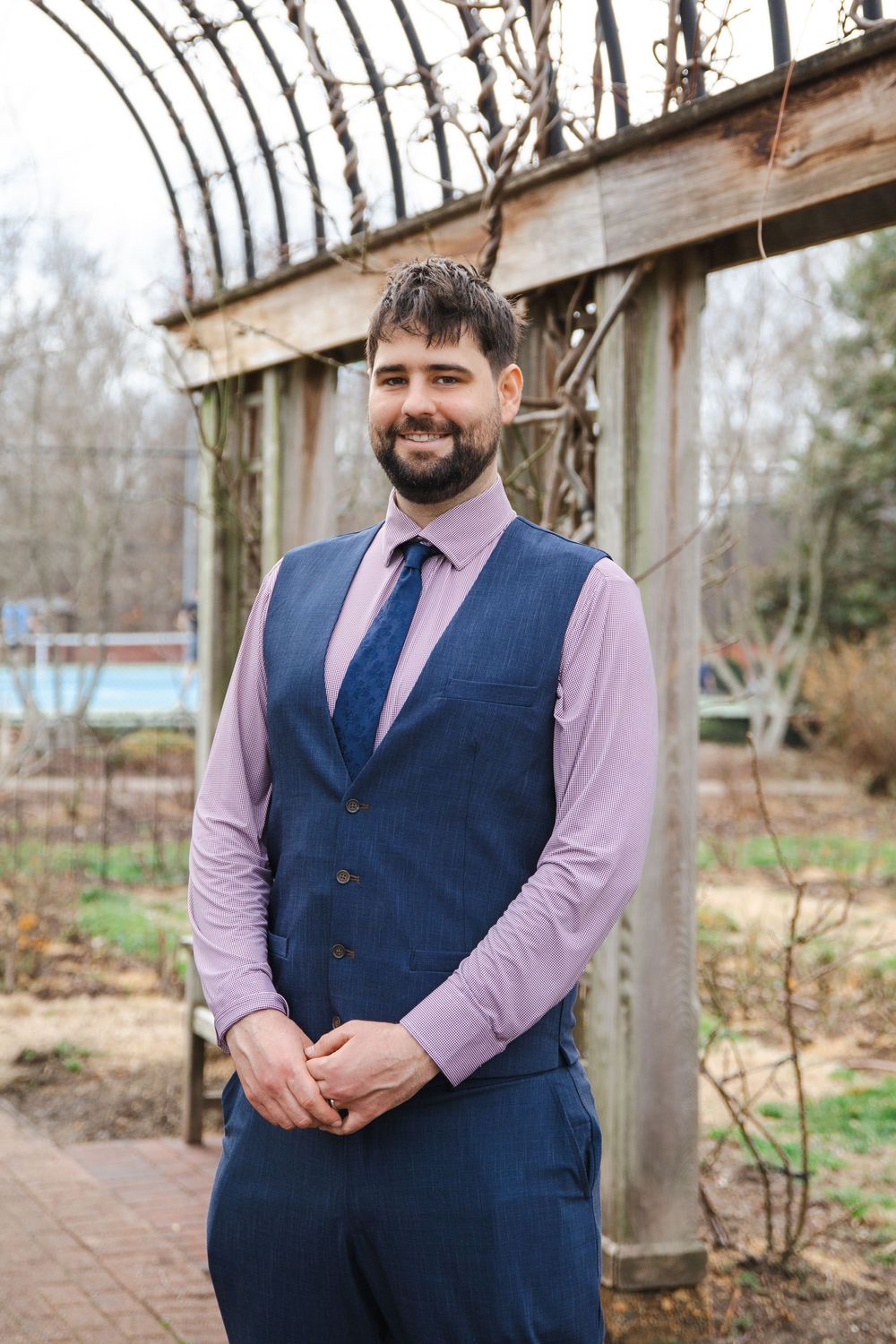 Smiling bearded man in blue vest, tie, and pink shirt standing outdoors under a wooden pergola.