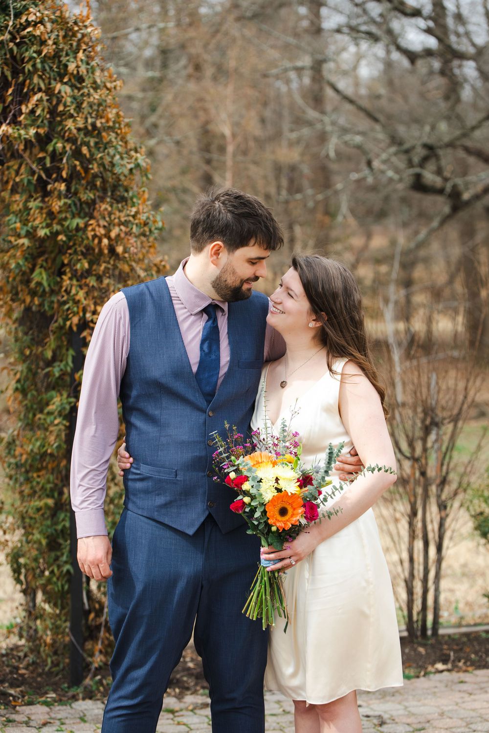A couple in wedding attire shares a romantic moment outdoors, the bride holding a colorful bouquet of sunflowers and wildflowers.