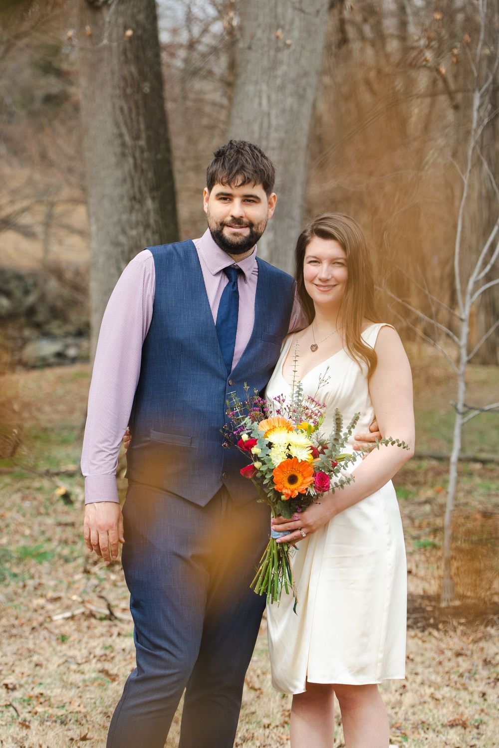 A smiling couple poses outdoors, groom in blue vest and tie, bride in white dress holding colorful flower bouquet.