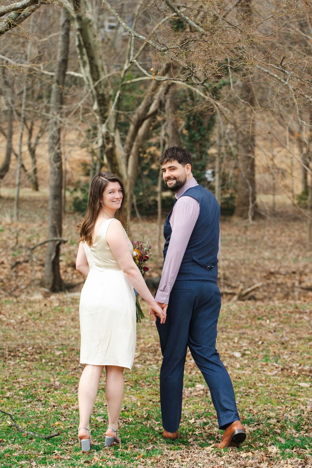 A couple holds hands outdoors in a park, woman in white dress with bouquet, man in navy suit, smiling during fall wedding.