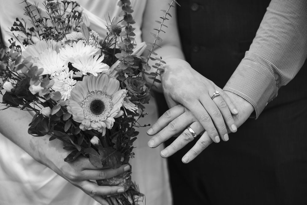 Black and white photo of bride and groom hands with wedding rings and bridal bouquet of flowers.