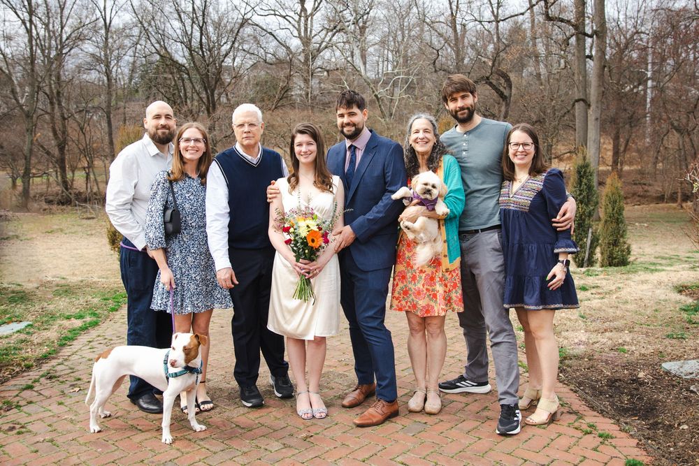 Wedding party poses outdoors in a park, bride holding bouquet, groom in navy suit, family smiling with a white dog.