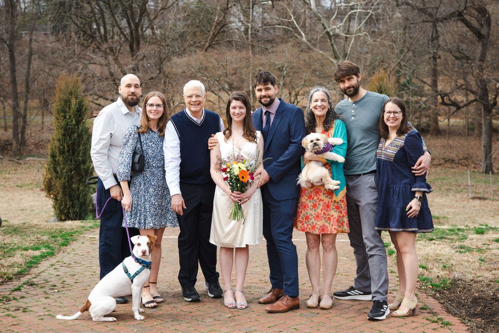 Family group photo at outdoor wedding with bride holding bouquet, groom, guests, and two dogs on a park path.