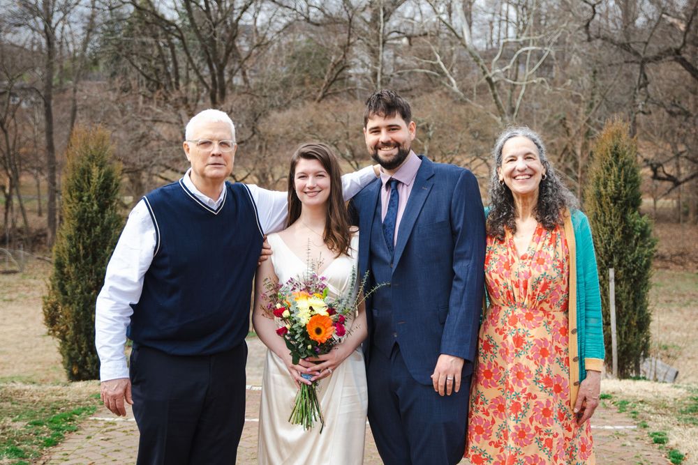 A bride and groom pose with family members outdoors at a wedding, surrounded by bare winter trees.
