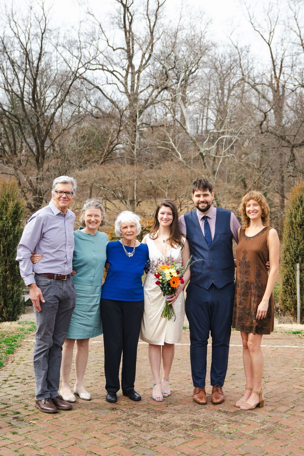 A wedding couple poses with family members outdoors on a brick path, bride holding colorful bouquet, bare trees in background.