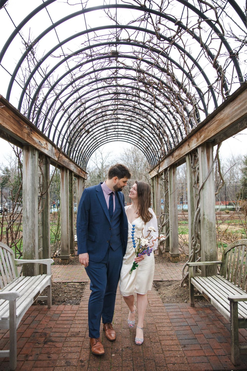Bride and groom share a romantic moment under a vine-covered pergola tunnel, bride holding a wildflower bouquet.