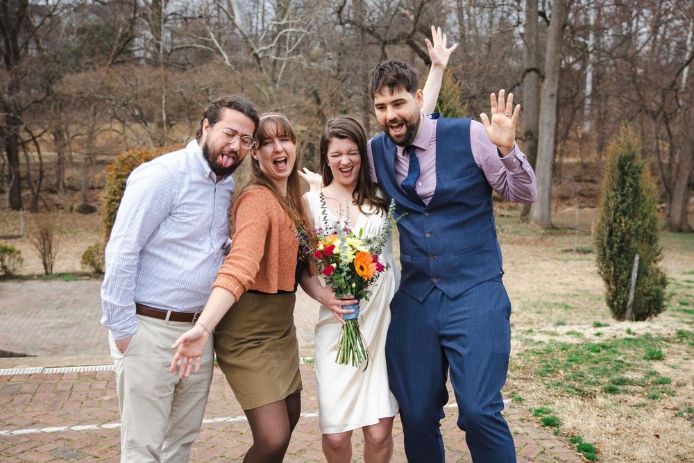 Joyful wedding party of four celebrating outdoors, bride holding colorful bouquet, groom in blue suit waving.