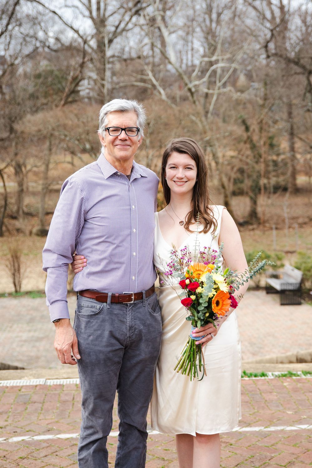 Older man and young woman in cream dress holding colorful bouquet posing together outdoors at a wedding ceremony.