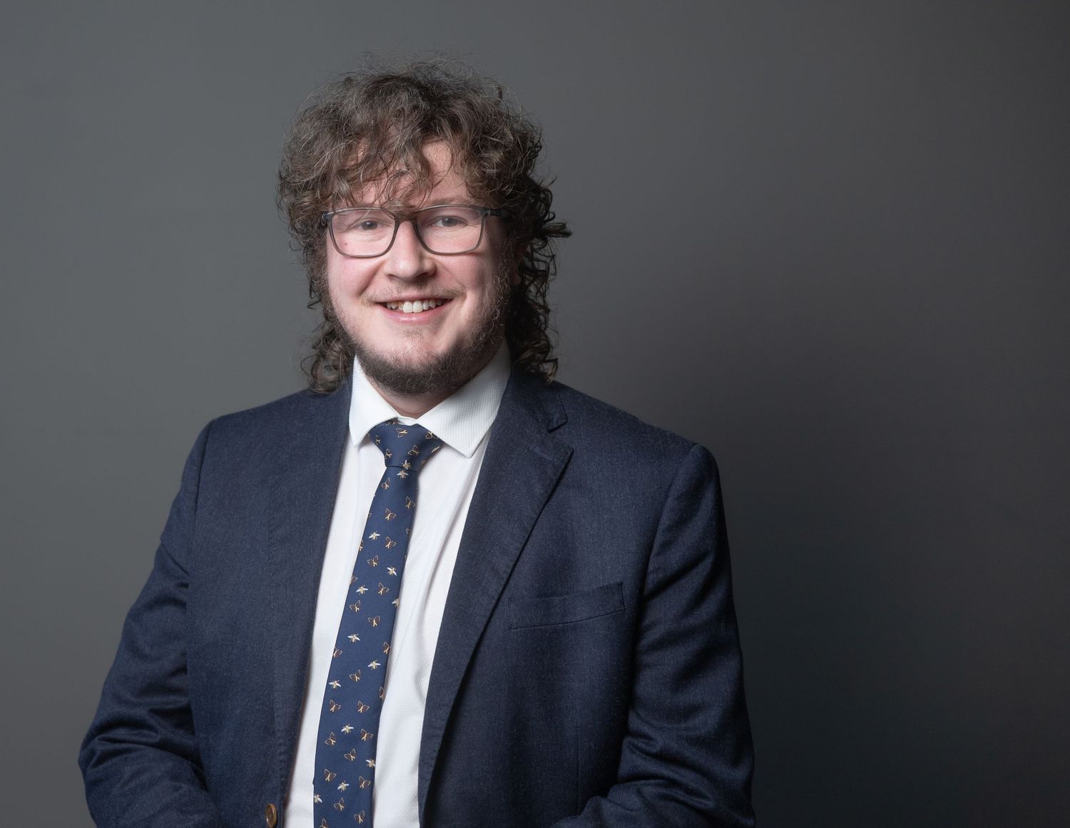 James in navy suit and patterned tie with curly hair and glasses, smiling against gray background.