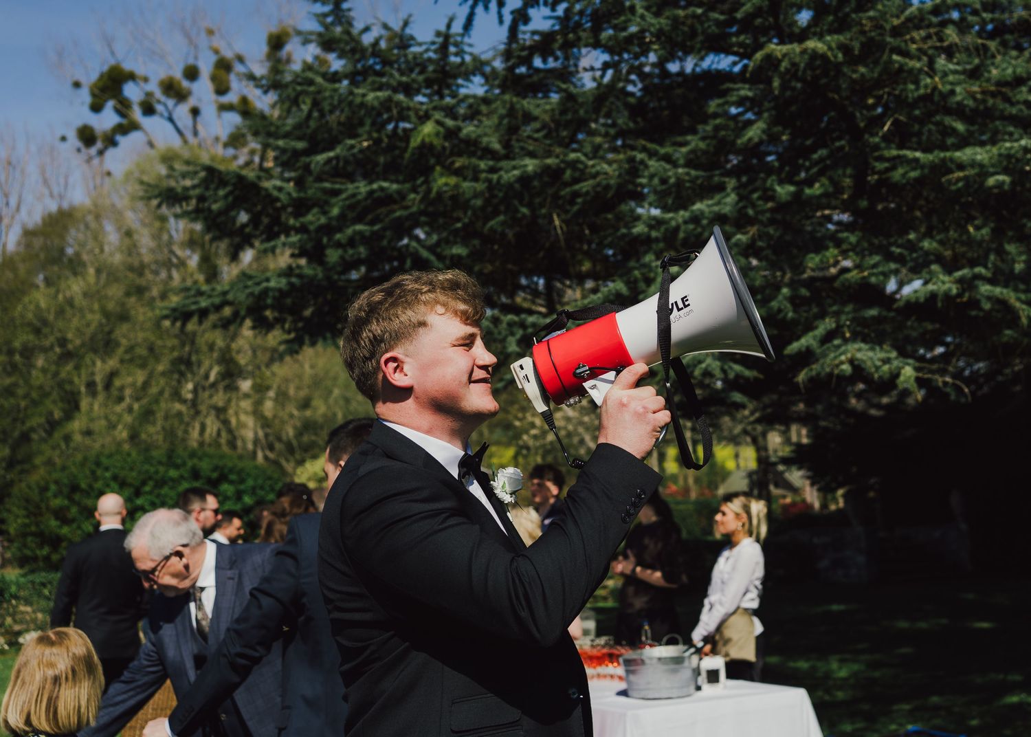 Man in suit speaking into red megaphone at outdoor wedding reception surrounded by guests and trees.