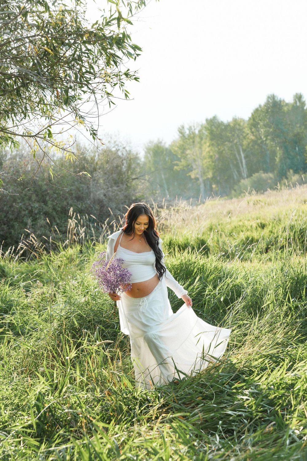 Pregnant woman in white dress holding lavender bouquet in sunny green meadow.