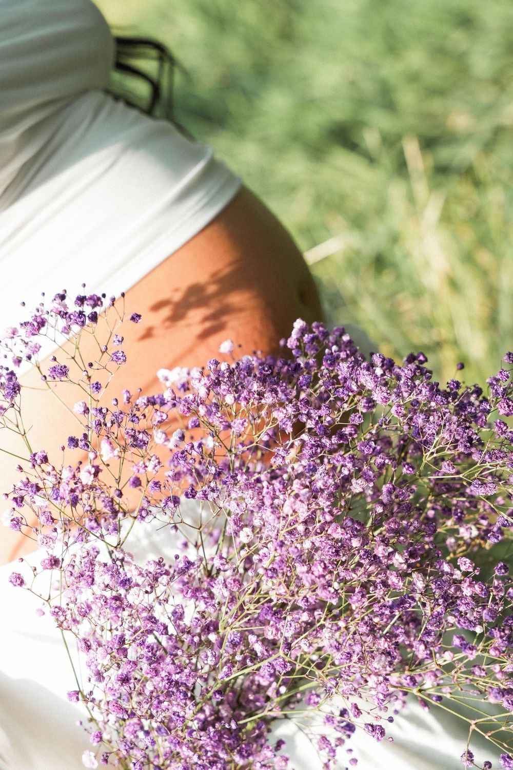 Person in white dress holding a bouquet of delicate purple wildflowers outdoors in a sunny green field.