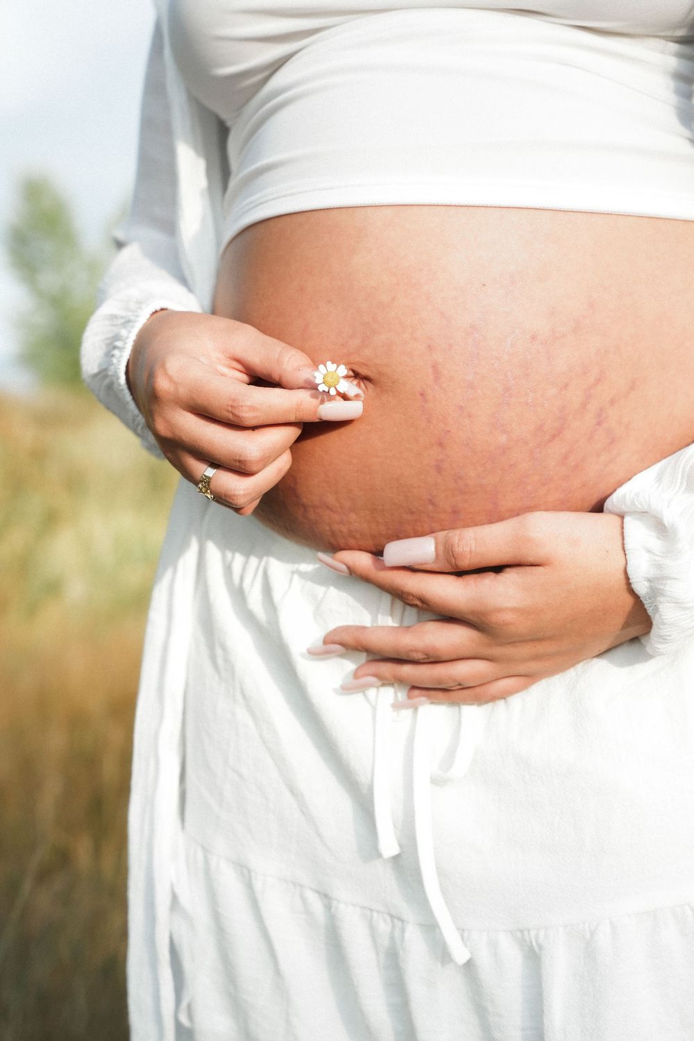Close-up of pregnant belly with stretch marks, woman in white outfit holding a daisy flower outdoors.