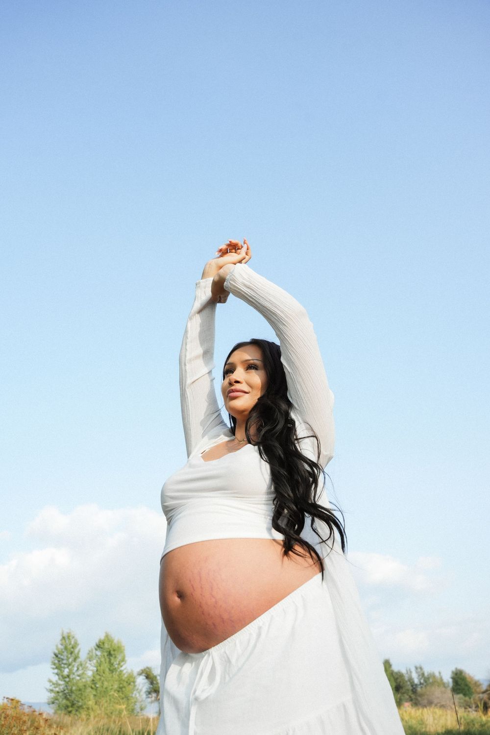 Pregnant woman in white outfit stretching arms overhead outdoors against blue sky, showing baby bump.