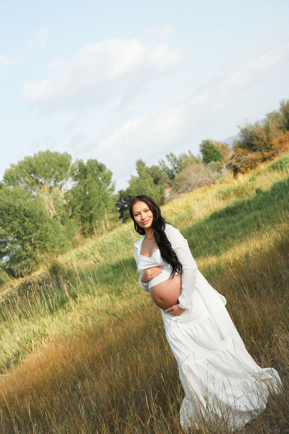 Pregnant woman in white dress holding belly in a scenic green field under a blue sky.