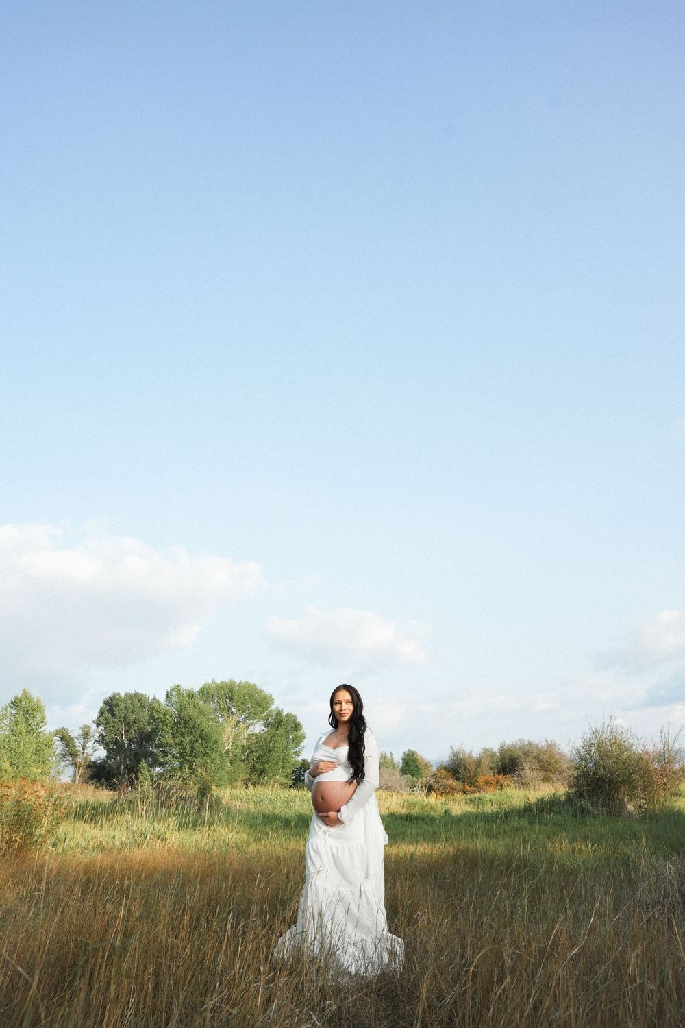 Pregnant woman in white dress standing in a green field, cradling her baby bump under a clear blue sky.