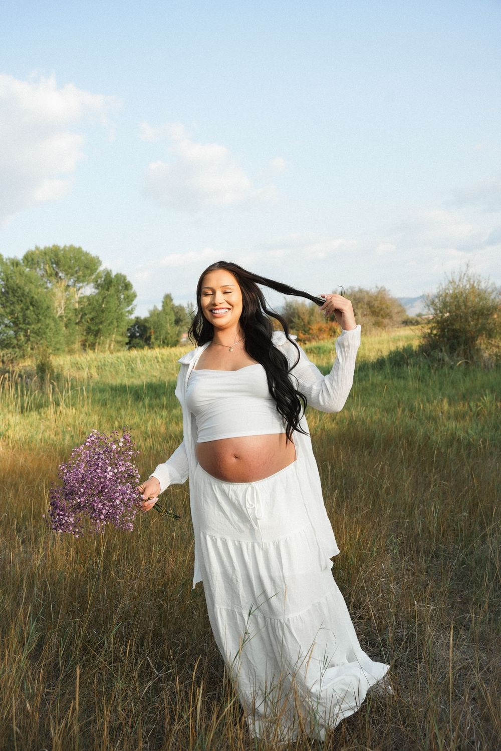 Smiling pregnant woman in white crop top and maxi skirt holding purple wildflowers in a sunny green field.