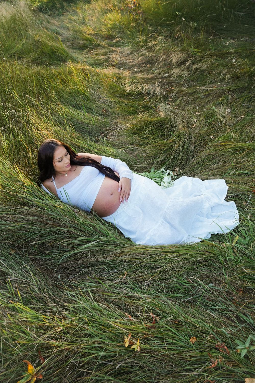 Pregnant woman in white dress lying peacefully in lush green grass field, cradling her baby bump outdoors.