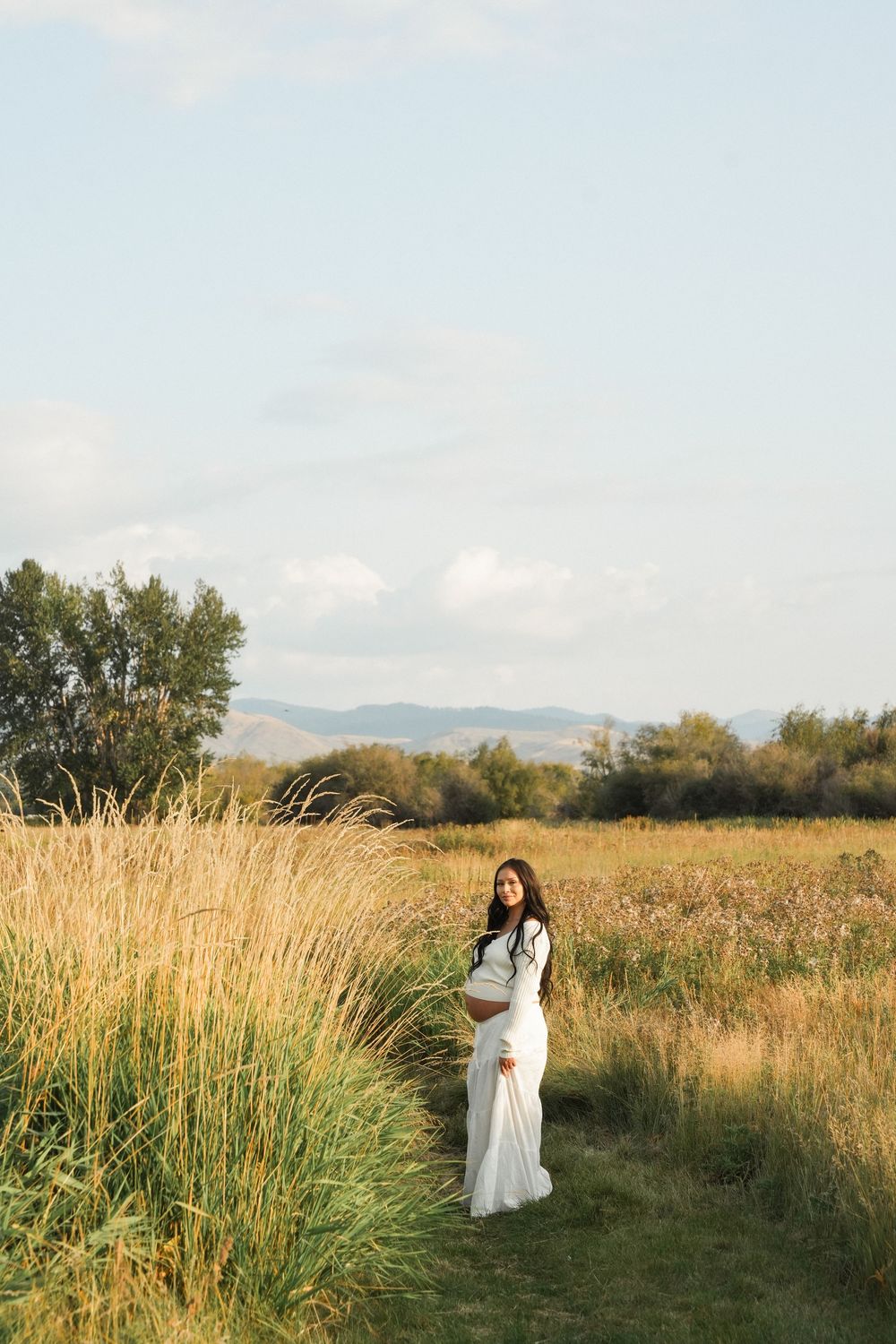 Pregnant woman in white dress standing in golden field with mountains and blue sky background.
