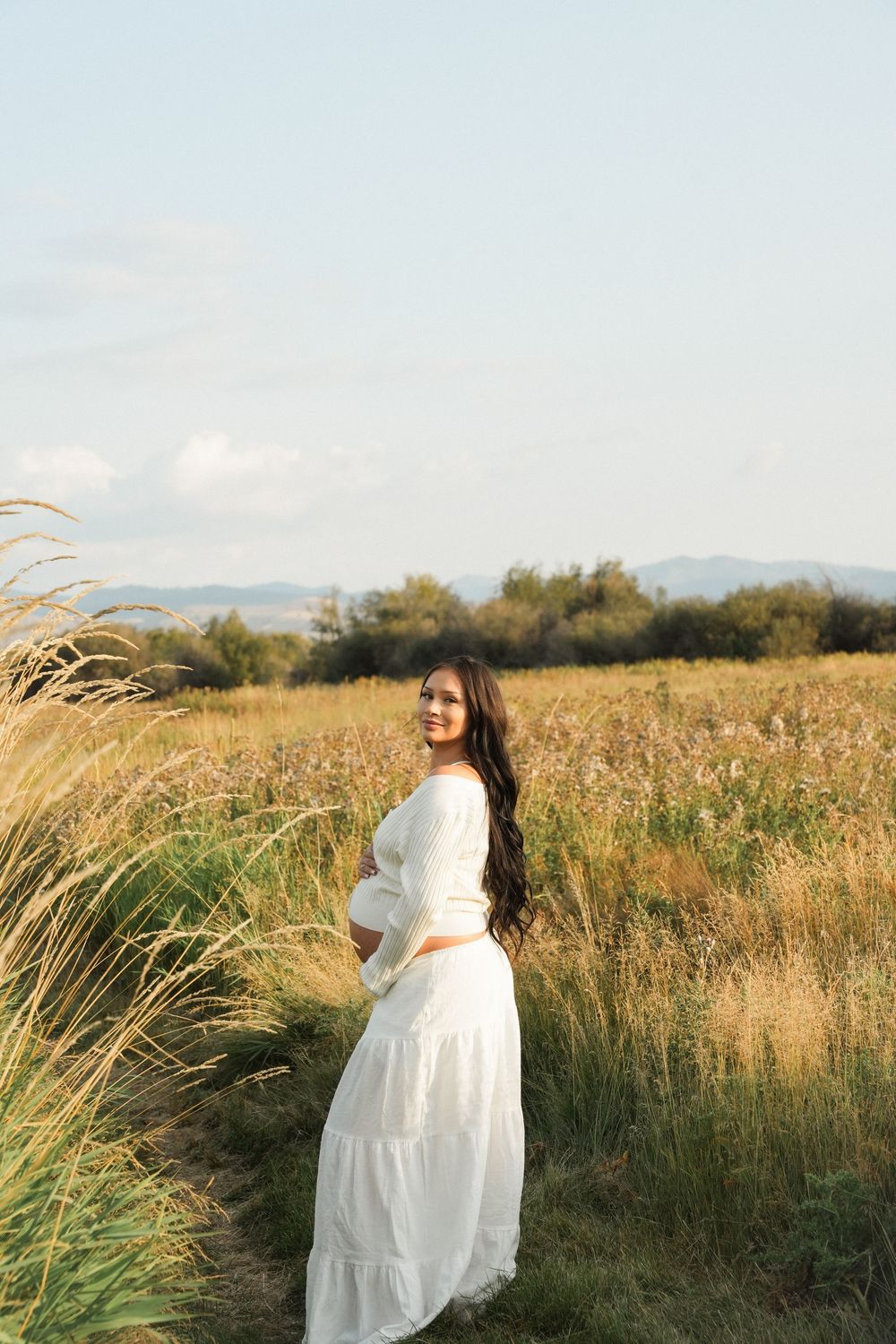 Pregnant woman in white dress standing in golden field, cradling baby bump, smiling over shoulder at sunset.