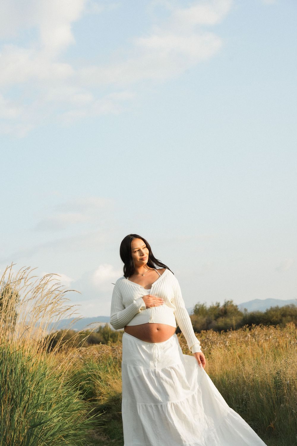 Pregnant woman in white crop top and skirt cradling baby bump in sunny outdoor field setting.