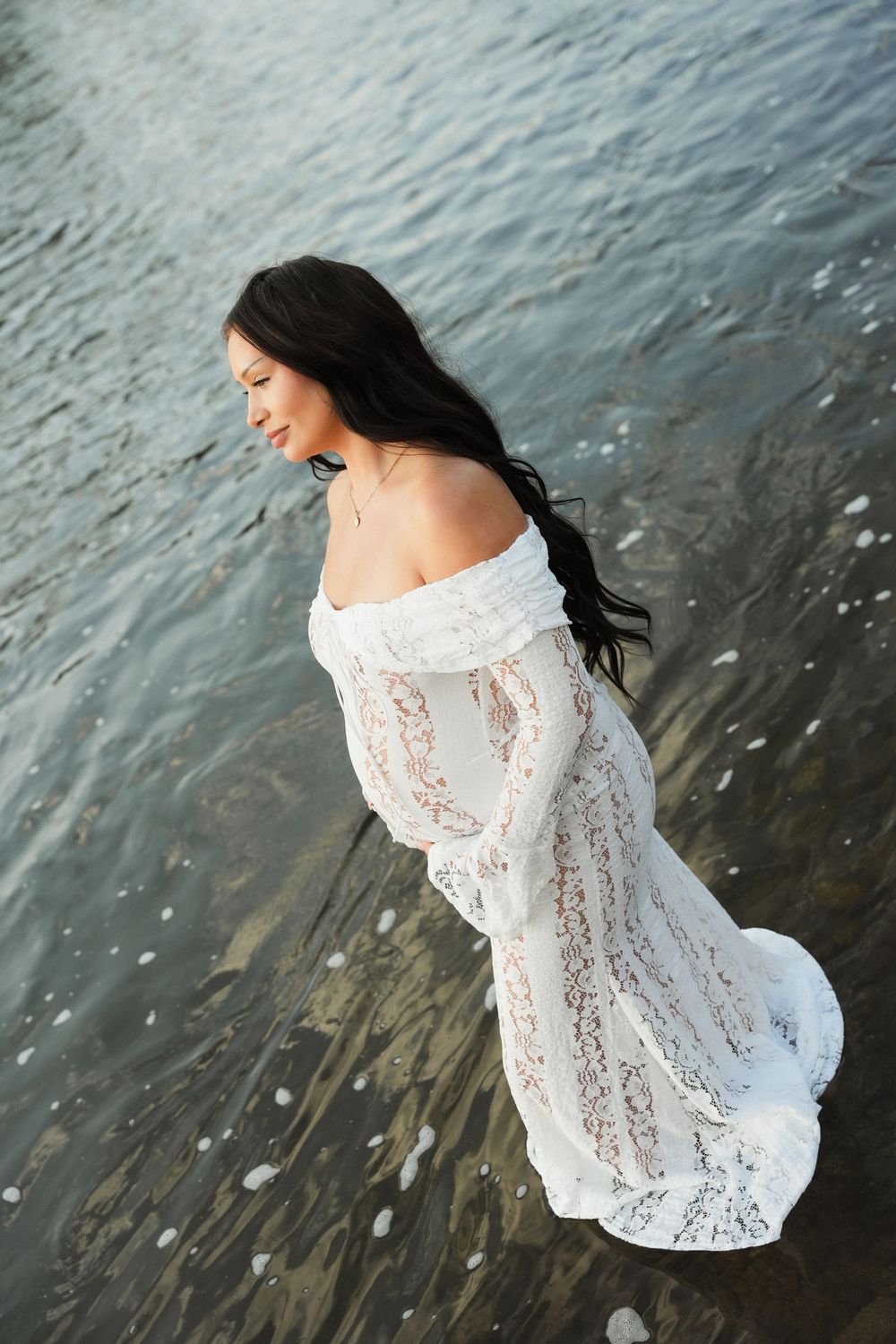 A woman in a white lace off-shoulder gown stands gracefully in shallow water with her dark hair flowing.