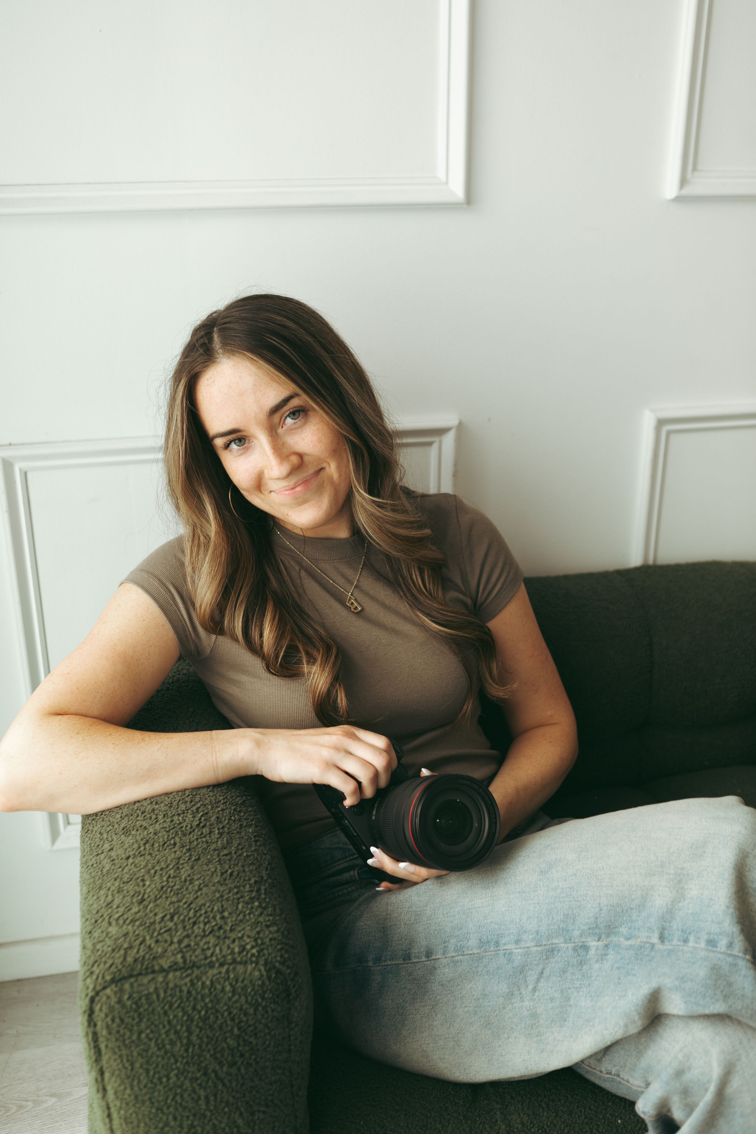 Young woman photographer smiling on green sofa holding DSLR camera in bright white room wearing casual outfit.