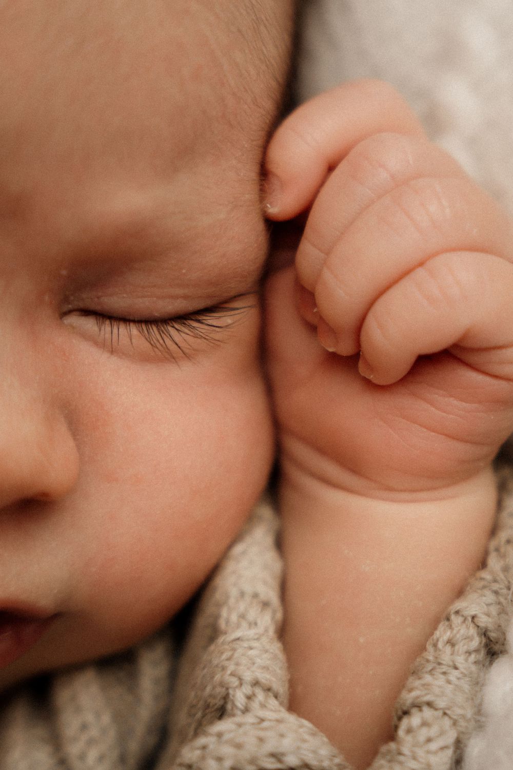Close-up of a sleeping newborn baby with tiny clenched fist beside face, wrapped in soft knit blanket.