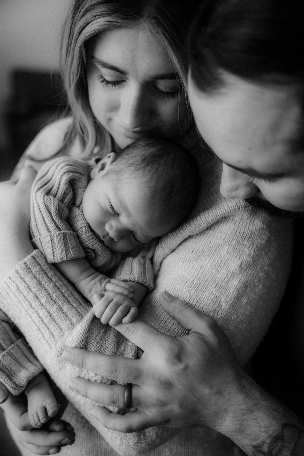 Black and white photo of parents tenderly kissing their sleeping newborn baby wrapped in a soft blanket.