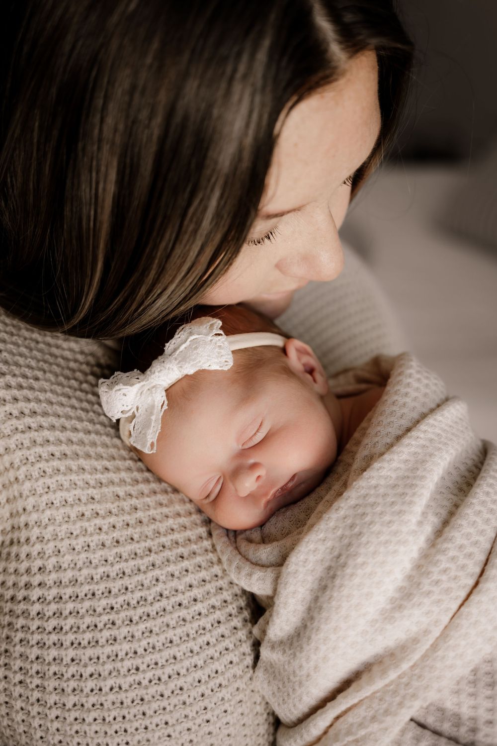 A mother tenderly holds her sleeping newborn baby girl wearing a white lace headband, wrapped in a soft beige blanket.