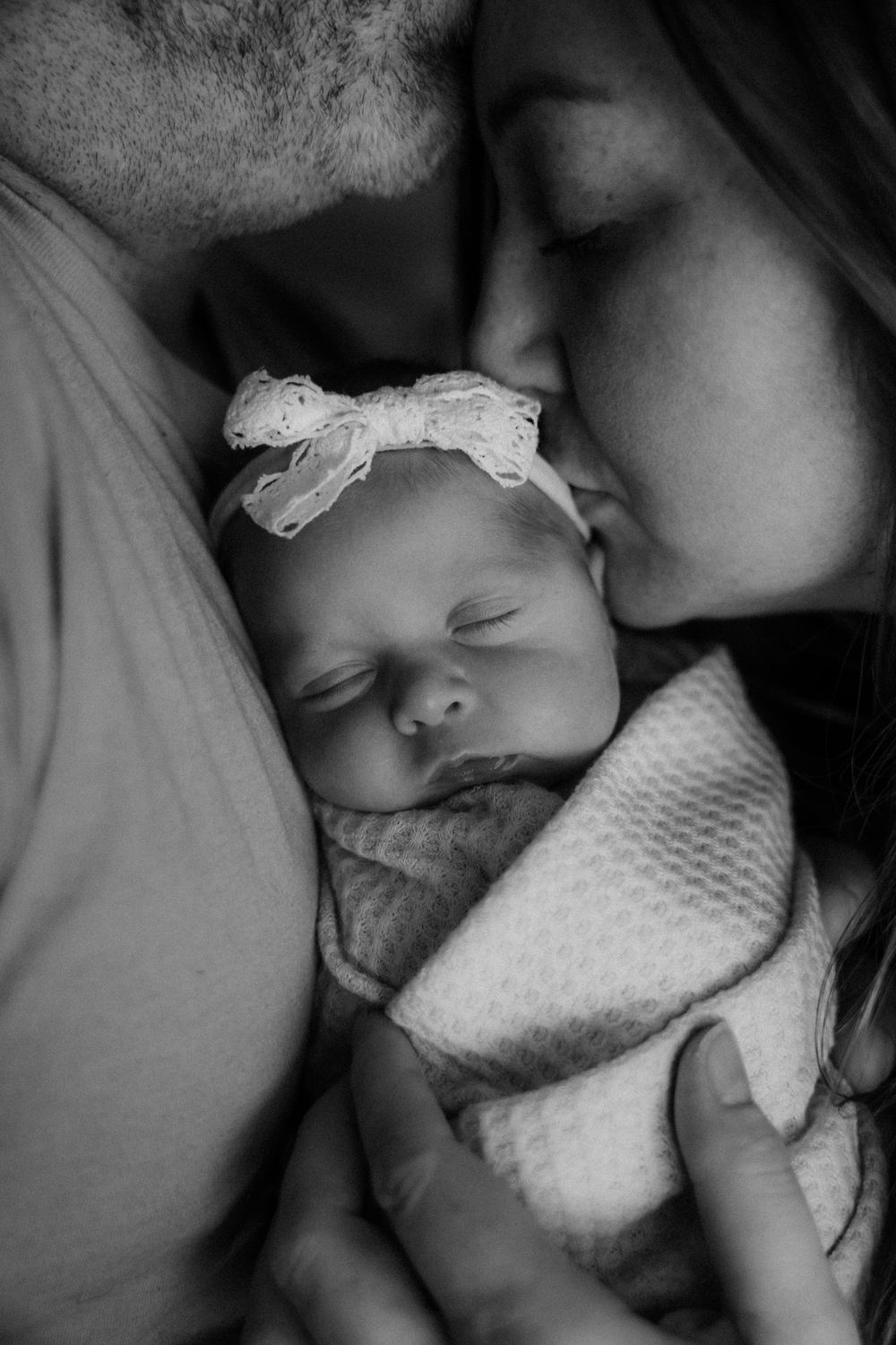 Black and white photo of parents kissing their sleeping newborn baby girl wearing a white lace headband.