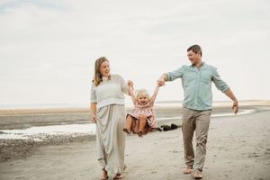 Family of three walking on beach, parents swinging toddler by hands, smiling and enjoying sunny day by the ocean.