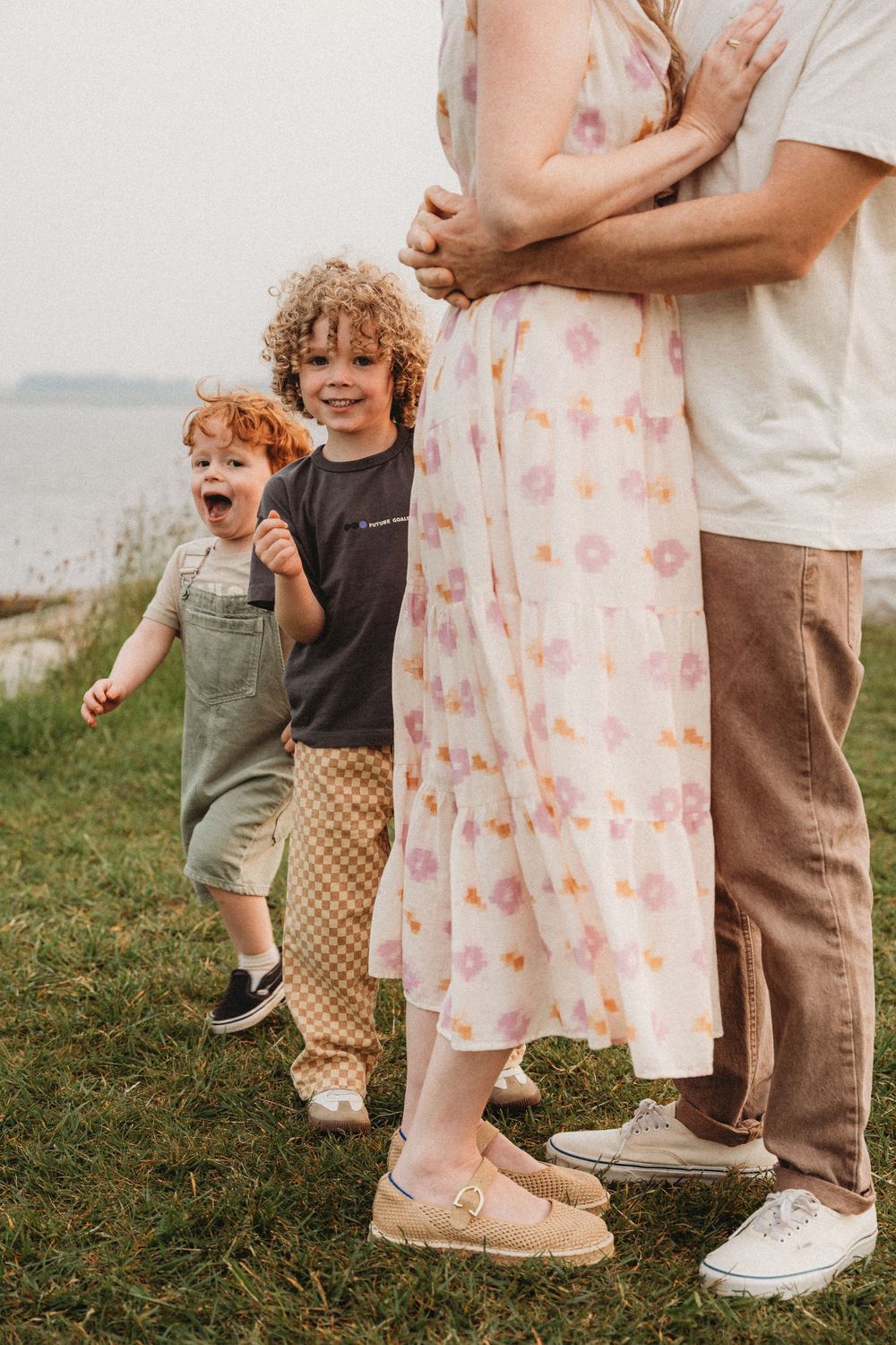 A family with two curly-haired boys standing outdoors near water, mother in floral dress embraced by partner.