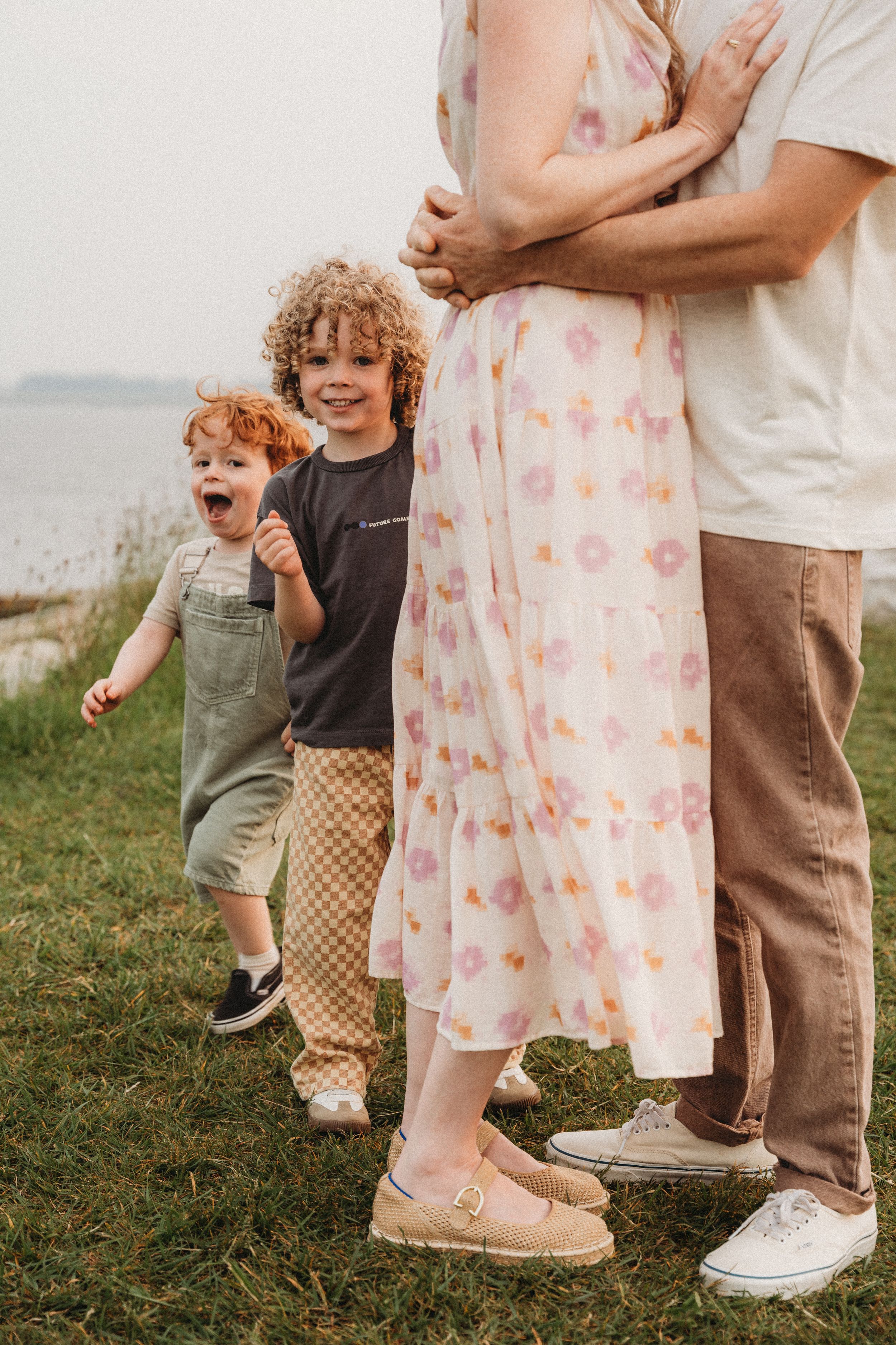 A family with two curly-haired boys standing outdoors near water, mother in floral dress embraced by partner.