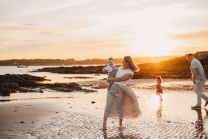 Family enjoying a golden sunset beach walk, mother holding baby while child plays in shallow water.