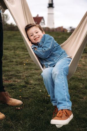 Happy young boy in denim jacket and jeans smiling in a hammock outdoors near a lighthouse.