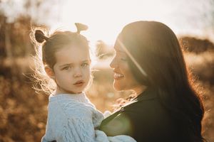 A mother holds her young daughter in golden sunset light, both wearing soft pastel outfits in an outdoor setting.